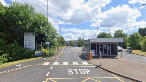 A Google street view image taken outside the entrance of the Moveero factory. There is a sign on the left in front of trees. And a barrier with "Stop" written in the road. 