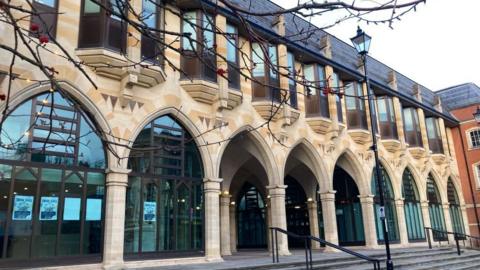The modern extension of the Guildhall in Northampton, showing a number of stone arches, with glass windows, part of a tree to the right, a lamppost and windows above. The building is light stone in colour. 