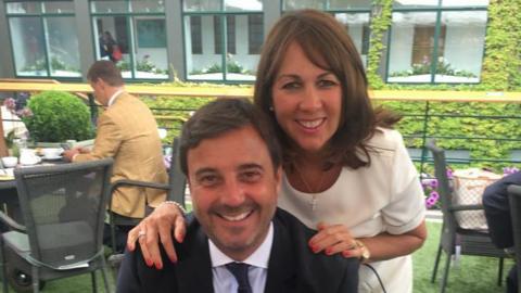 Jason and Gail O'Shea both smiling at the camera. Mr O'Shea is sitting in a chair in a cafe's outside dining area. Mrs O'Shea is standing behind him and has her hands on his shoulders. He is wearing a dark suit and tie. She is wearing a white dress.