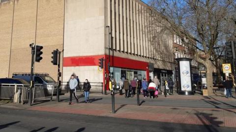 Exterior of the building - a tall cream-coloured building with a red entrance and people walking in front of it on the road. In the foreground is a traffic lighted pedestrian crossing.