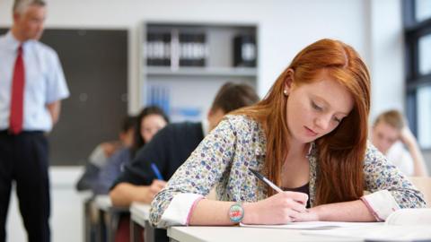 A girl with red hair is sitting in a classroom focusing on her work. She has a pen in her right hand, is wearing a flowery top and has a colourful wristwatch. There are other students behind her also focusing on their work and a teacher is watching over them.