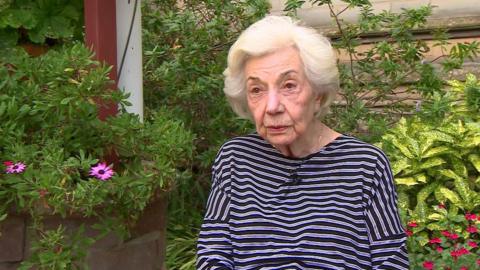 An elderly woman with white hair, wearing a navy and white striped top. She is sat outdoors in a garden.