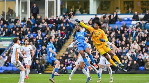 An action shot of Peterborough United's home clash against Shrewsbury Town. A goalkeeper in a yellow kit is flying through the air having just punched away the ball. Other football players - including some in blue kits and some in white kits - are watching the ball. Blurred football fans are in the background.