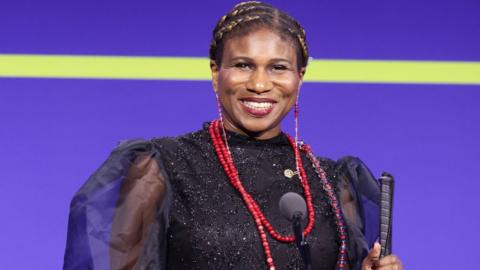 Lachi, a woman smiling in front of a purple screen with a yellow line going through it. Lachi is in front of a microphone and holding a cane with a leather handle.