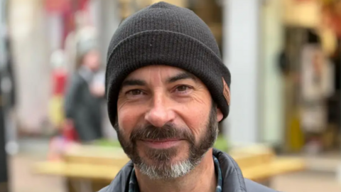 Mark Warren is smiling at the camera. He is wearing a black woolly beanie hat and has brown eyes and a brown and white beard. There are shops behind him.