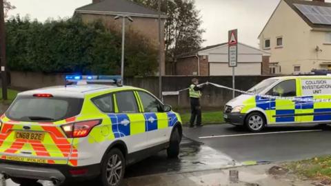 A police officer lifts a police cordon. To the left of the picture is a police car and on the right is a collision investigation unit van