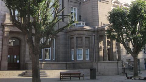 The entrance to the Jersey's Royal Court on a sunny day. In the paved area leading up to the door there are two trees, a bench and a bin. There are three steps leading up to the door which is brown wood and glass. The granite building is ornate with columns and elaborate block work.