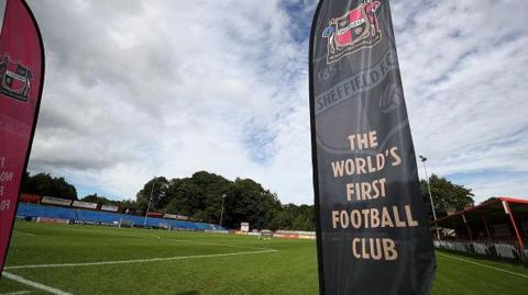 A football ground viewed from pitch level under a partly cloudy sky. In the foreground, a tall black banner stands upright near the touchline. The banner is branded with the Sheffield FC crest and the text “Sheffield FC – The World’s First Football Club”, printed in large, clear lettering.
The green grass of the football pitch stretches across the centre of the image, with white pitch markings visible near the edge. In the background to the right, a small covered stand with red seating is visible, along with perimeter advertising boards running along the far side of the pitch.