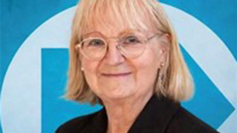 Sue Eacock. She has blonde hair and is wearing a dark jacket and standing against a blue Reform UK logo