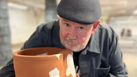 Johnny Vegas stands holding his ceramic artwork.