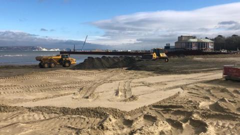 Sandy beach at Weymouth with the pavilion to the right hand side and see in the background - several diggers and dumpers are working on the beach.