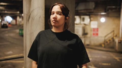 Ayse Hassan, standing in what appears to be an underground car park. She has quite short brown/black hair and is wearing all black. 