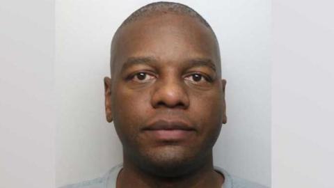 Mugshot of John Malonda, who has close-cropped hair, large brown eyes and is wearing a grey prison jumper.
