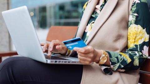A woman in a flowery blouse holds her bank card up to make a payment on a laptop