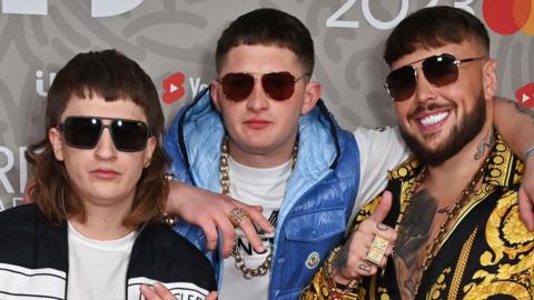 Bad Boy Chiller Crew's three members standing together in colourful outfits, gold chains and dark glasses on the red carpet at the Brit Awards in 2023