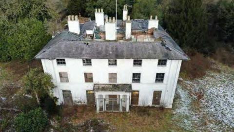 An aerial view of a derelict white building with severe roof damage, boarded windows and security fencing at the entrance. 