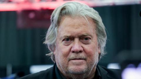 A close-up shot of Steve Bannon's face. He has long-ish grey hair and wears a black shirt.