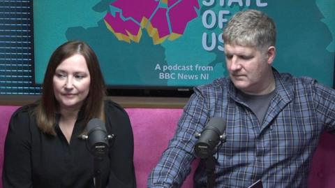 A screen shot of the Brysons filming a podcast. They are sitting in front of black microphones and in front of podcast promo posters. Ms Bryson has long brown hair and is wearing a black blouse. Mr Bryson has grey hair and is wearing a blue checked shirt.