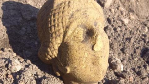 A stone Roman head carving lying on a pebbled ground. It is pale yellow and has its eyes closed and a damaged nose. 