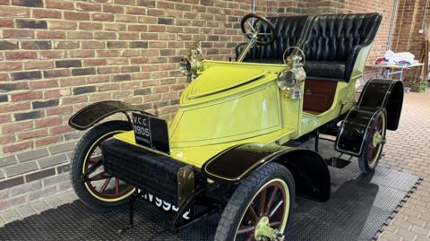 A vintage car with two raised leather seats and an exposed cabin. It is yellow and the wheels have yellow rims. A plate on the front reads: V.C.C 1905 Vauxhall. The car is parked in a building on rubber matting by a brown wall.