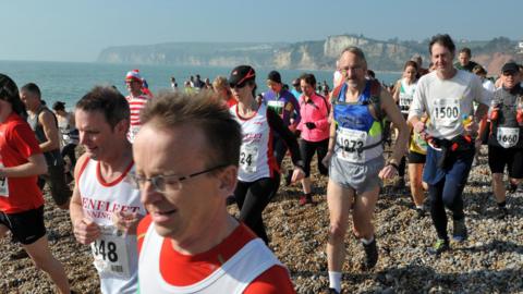 A large group of runners jog along Seaton beach in Devon as they take part in a race called The Grizzly. Cliffs and the sea can be seen in the background.