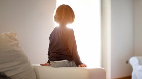 A boy sits with his back to the camera, with sun flooding through a window. He is sat on the arm rest of a sofa 