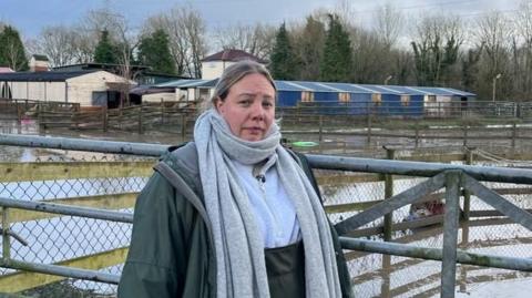 A woman wearing overalls, a jacket and a big scarf stands in front of a flooded field