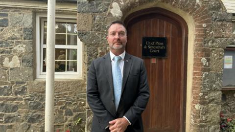 John Guille is pictured in a grey suit standing in front of a wooden door that has a black sign that says Chief Pleas & Seneschal's Court. There is a white flag pole next to him.