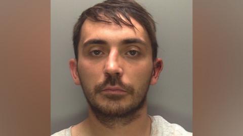 A police custody image of a man with dark hair, He is wearing a white top and he has a beard. He is looking directly at the camera.