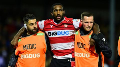 Josiah Edwards-Giraud holds his arms around two medics as he is helped off the field