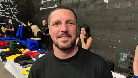 Tommy Coyle pictured at his school uniform giveaway during 2024. He has short black hair and a black beard and is wearing a black T-shirt. Behind him is a long table with various multi-coloured items of school uniform laid out. Five people can be seen behind the table who are working on handing out the items of clothing. 