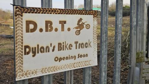 Metal corrigated fence with a sign labelled 'Dylan's Bike Track Opening Soon'. Through the fence is an open field. 