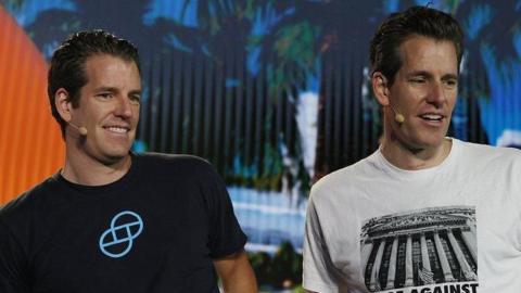 The Winklevoss twins: Two men. Both have dark hair and are standing and looking left. Both are wearing T-shirts. The shorter man on the left is wearing a black T-shirt with a blue logo on the front. The taller man on the right is wearing a white T-shirt marked with a black logo and the words "RAGE AGAINST THE MACHINE"