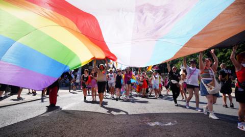 A big crowd of people in a Pride march walking and holding large rainbow flag above their heads.