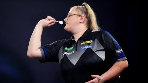 A woman with a long blonde pony tail wearing a black and blue darts shirt throws a dart