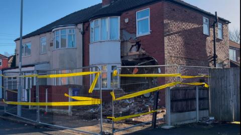 Fallen bricks lie on the ground in front of the severely damaged semi-detached house. There are further cracks and damage on the first floor, above the front door. Metal fences with yellow cordons surround the property.