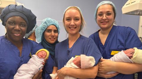 Four midwives wearing blue scrubs and hair nets. Three of them are holding a baby each. 