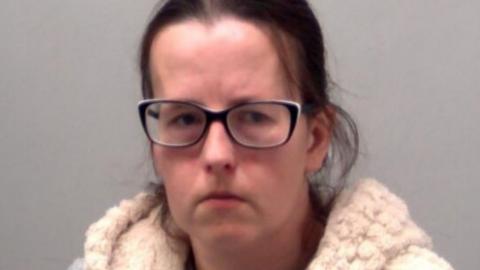 A police custody image of Emma Barnett. She has dark brown hair pulled back from her head and black framed glasses. She is wearing a crime gilet over a grey T-shirt. 