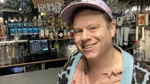 Mike is standing to the right of the picture, looking towards the camera smiling. He is wearing a blue and pink baseball cap, salmon pink colour top, and a blue and black cardigan. He is leaning against the bar in the club. 