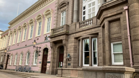 A picture of the outside of the Royal Court building in St Helier. There are two parts to the building, one is grey stone and the other is painted pink. 
