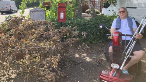 Sue Arnold wearing a white T-shirt and blue shorts- sitting on her red mobility scooter- next to the garden waste on the floor- in front of the red post-box