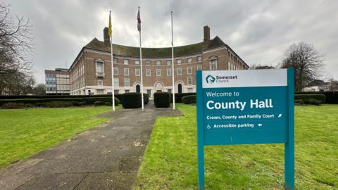 A turquoise sign reading "Welcome to County Hall" in the lawn outside a curved stone municipal building with flags flying on flagpoles outside.