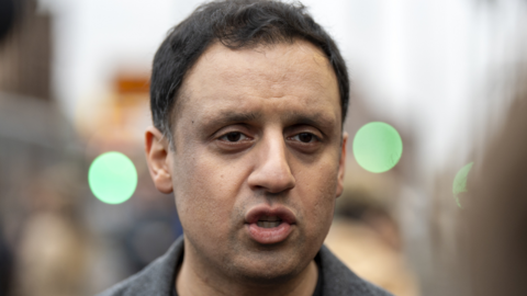 Anas Sarwar, with dark hair and wearing a grey jack, in a close-up shot with a blurred street background 