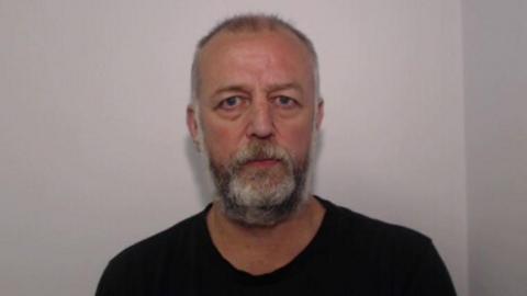 A mugshot of Andrew Talbot wearing a black shirt in front of a grey called wall.