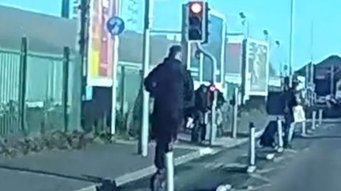 A man wearing dark clothing apppraoches a pedestial crossing with a red light riding an e-scooter