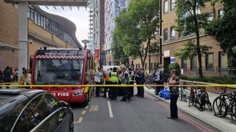 Emergency services are gathered by a fire vehicle outside a modern hospital building. There is tape closing off the road. 