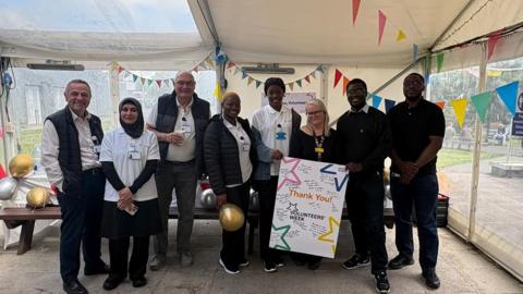 A group of people stand together inside a decorated tent, holding a “Thank You Volunteers Week” sign with bunting and balloons around them.
