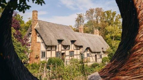 Anne Hathaway's cottage has creamy yellow stonework, timber framing and a brown thatched roof. It has three chimneys and is surrounded by trees and shrubbery to the front.