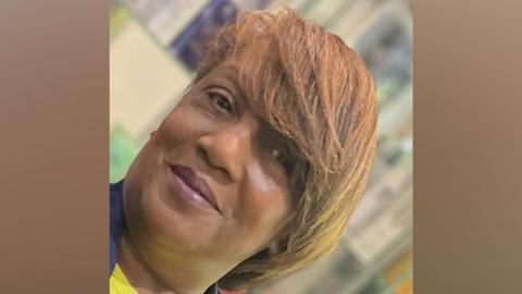 Close-up portrait of a person with short, side-swept light brown hair, looking toward the camera. The image is slightly tilted and framed tightly around the face and shoulders. The background is softly blurred, with indistinct indoor features visible behind the subject.