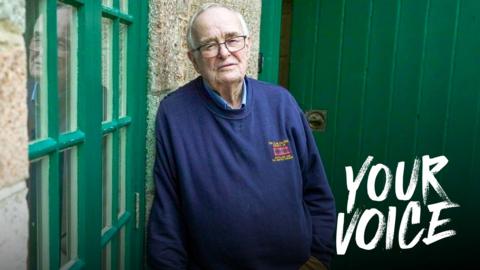 An older man with grey receding hair and glasses leans against a granite wall looking towards the camera. He wears khaki, flannel trousers and a blue sweatshirt. He looks sad.